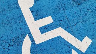 Blue background with a crackle surface like a paved parking spot painted with white international symbol for accessibility – the profile view of a stick figure in a wheelchair. 